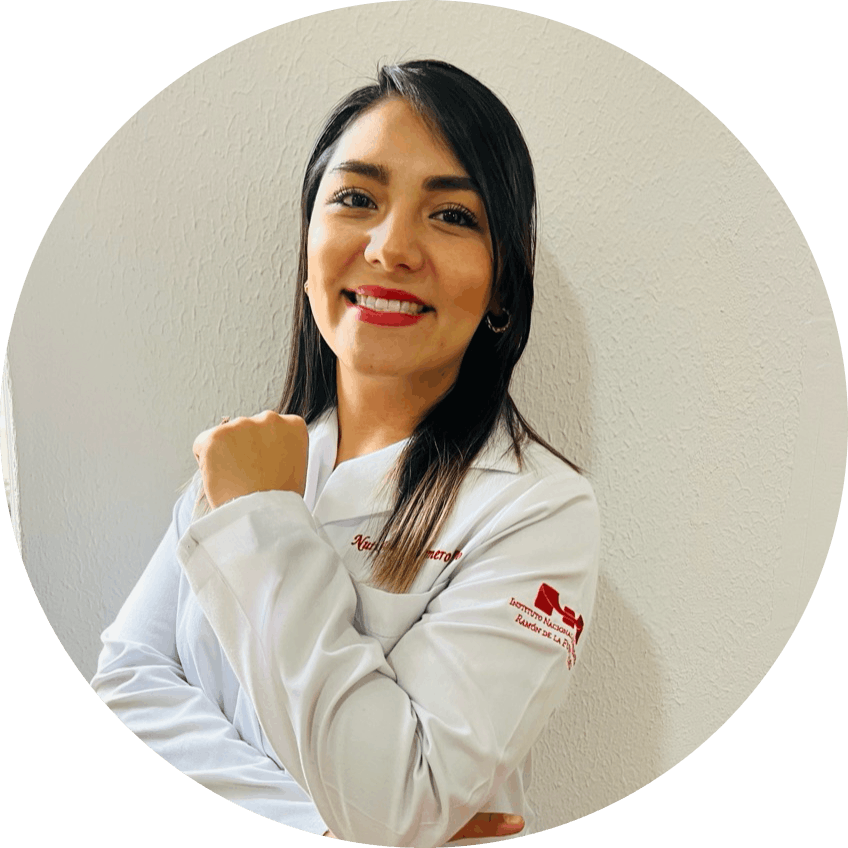 Smiling woman in a white lab coat with a logo, arms crossed, standing against a beige wall.